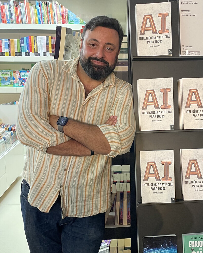David Carvalhão leaning against a bookshelf displaying copies of his book